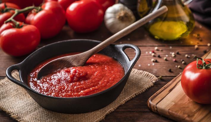 fotografia em tons de marrom e vermelho de uma bancada marrom vista de cima. Contém uma panela preta com molho de tomate e uma colher de madeira para mexer, ao lado esquerdo tomates.