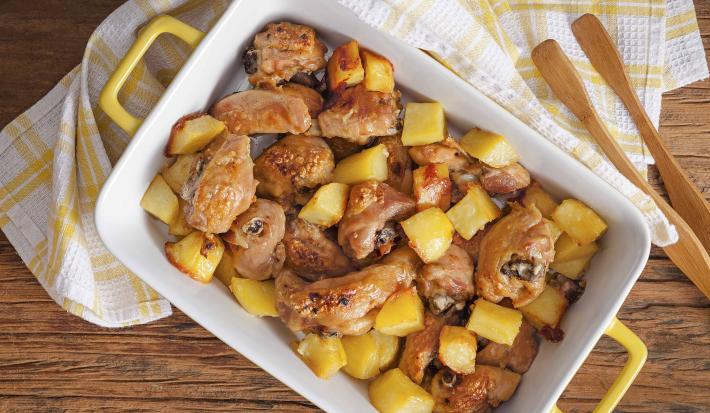 Fotografia em tons de branco e amarelo de uma bancada de madeira com um pano quadriculado branco e amarelo, sobre ele uma travessa com alças amarela por fora e branca por dentro com os pedaços de frango e batata, ao lado um pegador de madeira.
