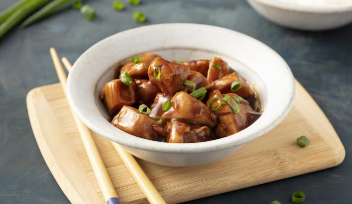 Fotografia em tons de marrom em uma bancada de madeira cinza escura com uma tábua de madeira ao centro, com um prato redondo fundo bege e o frango teriyaki com cebolinhas verdes dentro dele. Ao lado, hashis apoiados na tábua.
