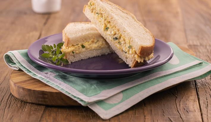 Foto em tons de bege da receita de pãozinho com ovo mexido servida em um prato de porcelana roxo, sendo duas metades do sanduíche, uma por cima da outra. O prato se encontra sobre uma tábua de madeira com um paninho verde claro entre eles.
