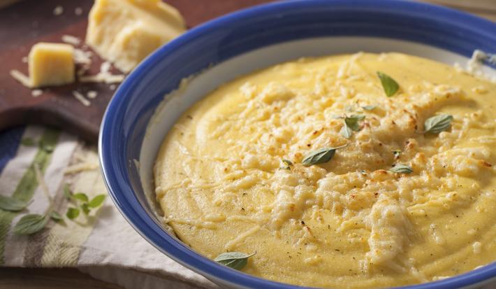Fotografia em tons de marrom e azul de uma bancada marrom com um paninho branco, sobre ele um recipiente redondo branco e azul com polenta. Ao fundo uma tábua com queijo parmesão.