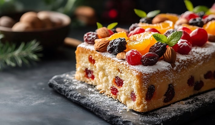 Fotografia de uma fatia de bolo retangular de Natal decorado com frutas secas, castanhas e folhas de hortelã, polvilhado com açúcar de confeiteiro e apresentado em uma base de pedra escura.
