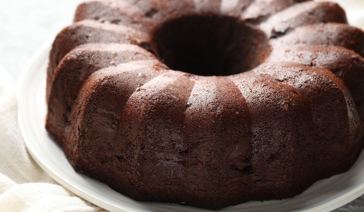 Bolo de chocolate bundt, com cor marrom escuro, repousa em um prato branco. A superfície brilhante e as ranhuras verticais criam um padrão atraente.