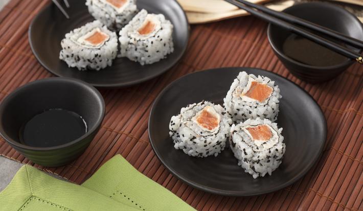 Foto em tons de marrom da receita de uramaki de salmão servida em 6 porções divididas em dois pratos pretos com um recipiente com shoyo ao lado, além de hashis pretos também. Tudo isso sobre uma esteira marrom escura com um paninho verde à frente