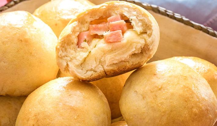 Fotografia em tons de amarelo em uma bancada de madeira vermelha um pano azul, uma cesta de vime com pães recheados com queijo.