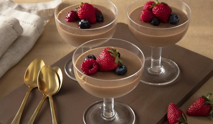 Foto em tons de marrom da receita de mousse de chandelle com frutas vermelhas servida em três taças de vidro sobre uma base marrom escuro com três colheres douradas ao lado, além de um paninho bege e morangos