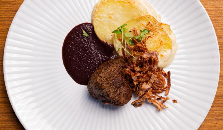 Fotografia vista de cima de um prato branco raso com molho de vinho tinto, cebolas fritas, filé mignon e batatas gratinadas. O prato está sobre uma bancada de madeira clara.