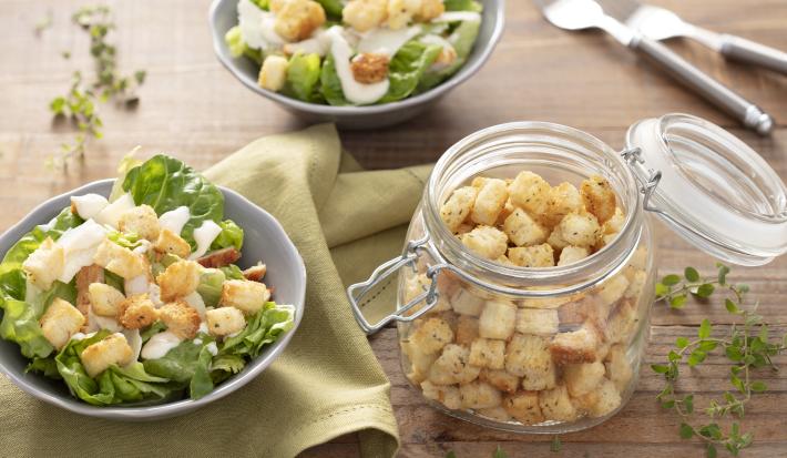 Fotografia em tons de verde em uma bancada de madeira clara, um pano verde, dois bowl com os croutons com a salada de alface e molho de iogurte. Ao centro, um pote de vidro hermético com vários croutons dentro dele.