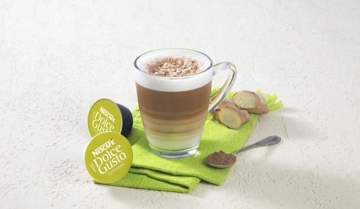Fotografia em tons de verde em uma bancada de madeira clara com um pano verde e uma xícara de vidro com o cappuccino dentro dele. Ao lado, cápsulas de Dolce Gusto Cappuccino.
