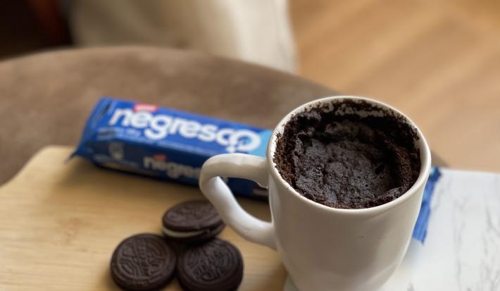 Foto de uma caneca branca contendo a receita de Bolo Negresco, sobre uma tábua com alguns Biscoitos Negresco soltos e um pacote em tom azul de Biscoito Negresco