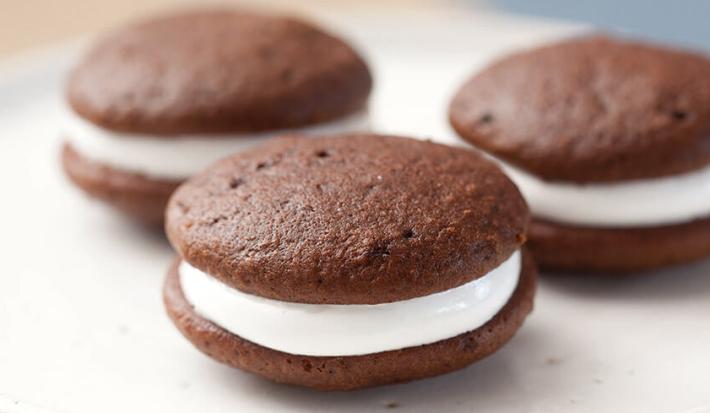 Fotografia em tons de preto em uma bancada de madeira clara, um prato redondo branco raso com três sanduicinhos de chocolate recheados com marshmallow.
