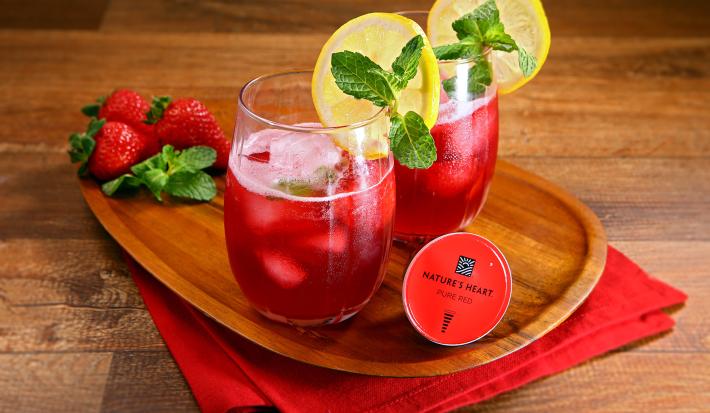 Fotografia em tons de vermelho em uma bancada de madeira clara, um paninho vermelho, uma tábua de madeira e dois copos de vidro com o chá de limão com frutas vermelhas. Ao lado, morangos e a cápsula do chá de Pure Red Nature´s Heart.