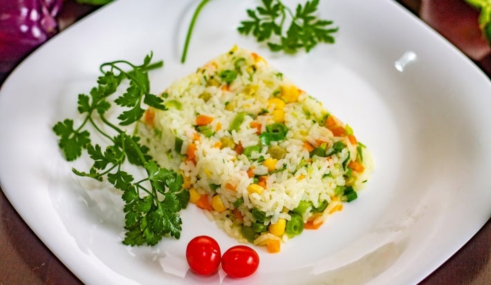Em um prato de porcelana branco, há um porção colorida de arroz amarelo, milho verde,  ervilha, decorados com tomate cereja e galhos de coentro.