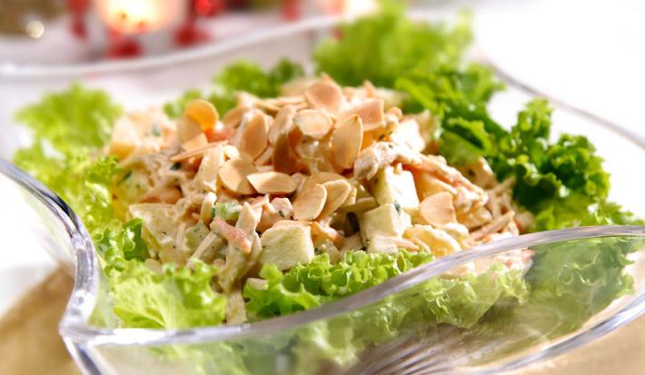 Fotografia em tons de verde em uma mesa com uma toalha branca, um apoiador de prato de madeira, um recipiente de vidro fundo com a salada de alface e frango dentro dele.