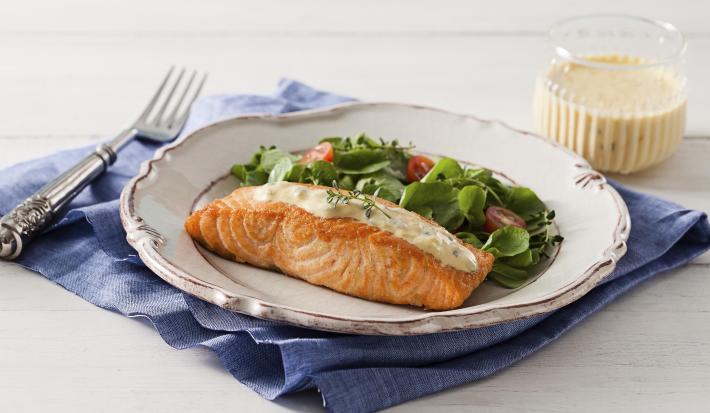 fotografia em tons de branco e azul de uma bancada branca vista de frente, contém um pano azul, um prato redondo branco com um pedaço de salmão com creme branco por cima e folhas com fatias de tomate cereja para acompanhar. Ao lado um garfo para servir