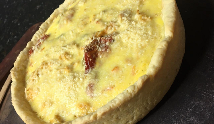 Foto em tons de amarelo da receita de quiche de frango com tomates secos servida em uma porção sobre uma mesa de pedra preta bem escura