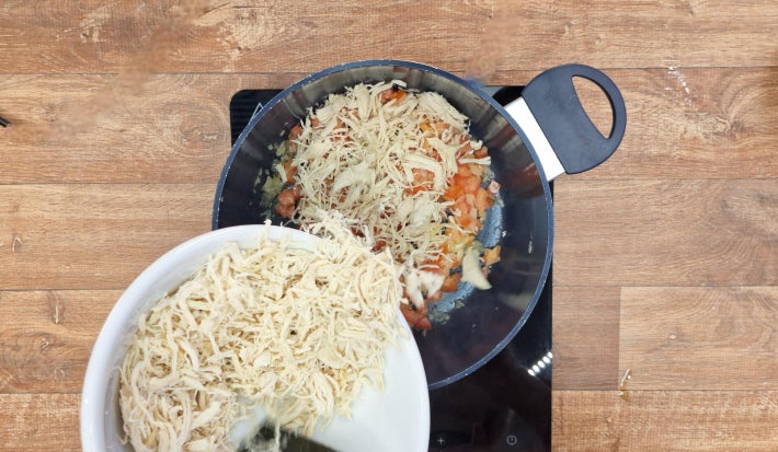 Frango desfiado sendo despejado de tigela dentro de panela com tomates e cebolas refogando sobre fogão preto portátil