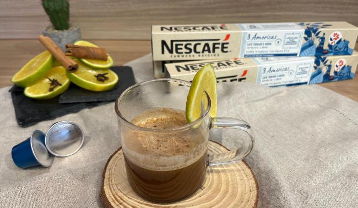 Fotografia em tons de amarelo e azul com uma xícara de vidro ao centro. Dentro da xícara, que está enfeitada com uma fatia de laranja,  existe uma mistura de café com suco de laranja. Ao fundo existe fatias de laranja e duas caixas de cápsulas NESCAFÉ