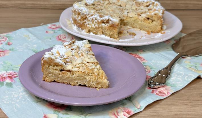 Fotografia em tons de lilás com um prato lilás ao centro. Em cima do prato existe uma receita de bolo de cor caramelo feito com farinha de amêndoas torradas.
