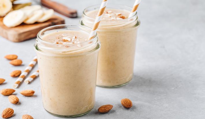 Fotografia em tons claros de uma mesa de cimento queimado claro. Sobre a mesa tem dois copos de vidro com milkshake de coco e amêndoas, com lascas de amêndoas sobre cada um. Dentro de cada copo tem um canudo branco e marrom, ao lado de amêndoas.