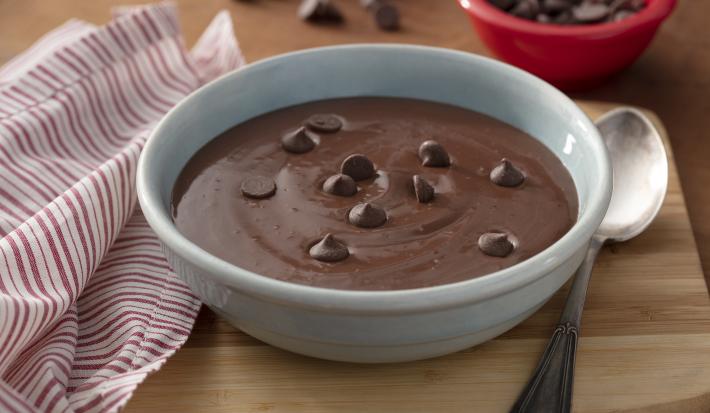 Fotografia em tons de vermelho em uma bancada de madeira clara, um pano branco com listras vermelhas, uma tábua de madeira com um recipiente redondo fundo com a sobremesa mingau de chocolate dentro dele e algumas gotas de chocolate para decorar.