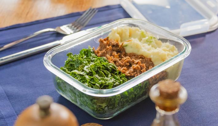 A fotografia mostra uma marmita em pote de vidro com carne desfiada ao meio, purê de batata-doce cremoso no canto superior direito e couve refogada verde no canto esquerdo.
