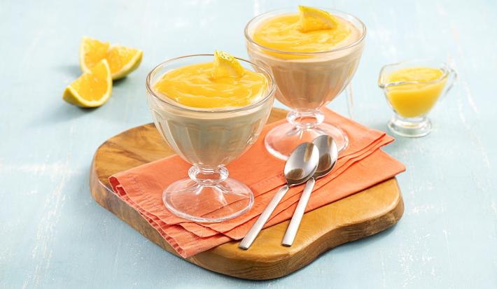 Foto da receita de Mousse de Cappuccino com Calda de Laranja. Oberva-se duas taças transparentes com a mousse e a calda laranja por cima em cima de um guardanapo laranja e uma tábua redonda de madeira. Como decoração, pedaços de laranja em um fundo azul.