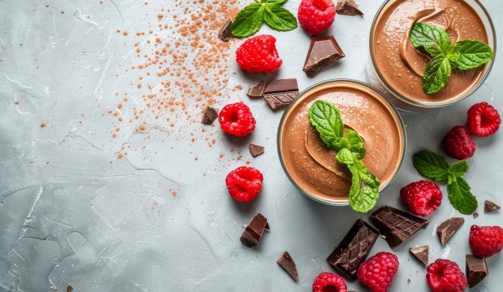 Fotografia vista de cima de dois potes de vidro sobre uma mesa de cimento queimado. Dentro do copos tem a sobremesa, finalizada com ganache e folhas de hortelã. Ao redor dos copos tem hortelãs, pedaços de chocolate e framboesas espalhadas.