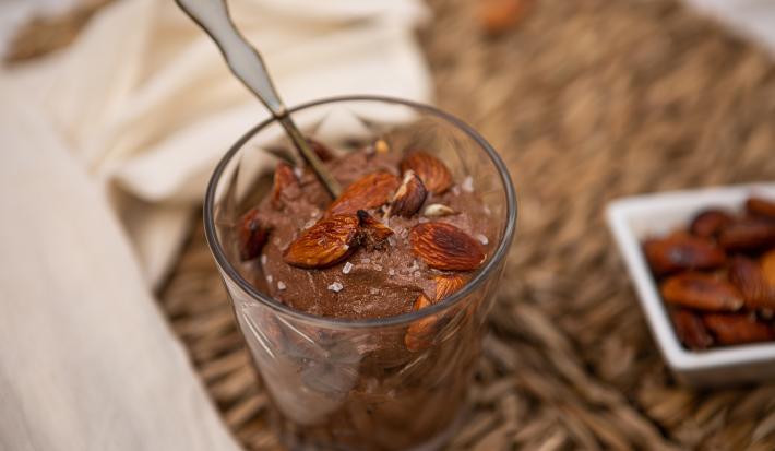 Fotografia de uma mistura de chocolate meio amargo, nozes e creme de leite em um recipiente de vidro. Por cima, nozes, flor de sal e uma colher sobre o creme. Ao lado, um pano branco e um recipiente branco de vidro com nozes.