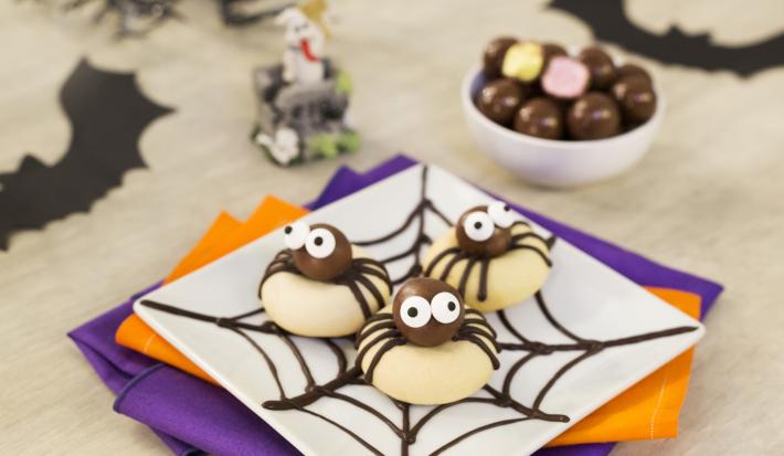 Imagem aproximada de um prato quadrado branco com três biscoitos em formatos de aranhas. A bancada está decora com papéis em tons roxo e laranja e alguns morcegos de mentira.