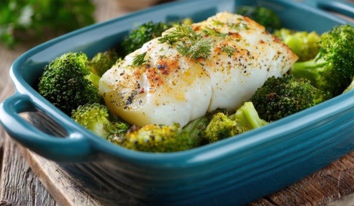 Em cima de uma mesa, refratário azul escuro com posta de bacalhau e floretes de brócolis.