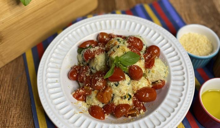 Fotografia em tons de amarelo e vermelho em uma bancada de madeira com um pano colorido em azul, vermelho e amarelo e um prato branco fundo em cima dele com o gnudi e molho de tomate por cima dentro do prato. Ao lado, potinhos com queijo ralado e azeite.