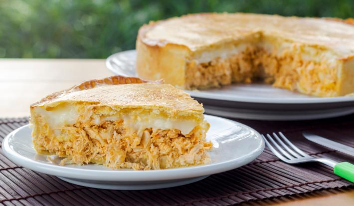 Fotografia de uma fatia de torta de frango com requeijão sobre um prato pequeno de vidro raso. No fundo tem um apoio branco e preto com o restante da torta quase inteira.
