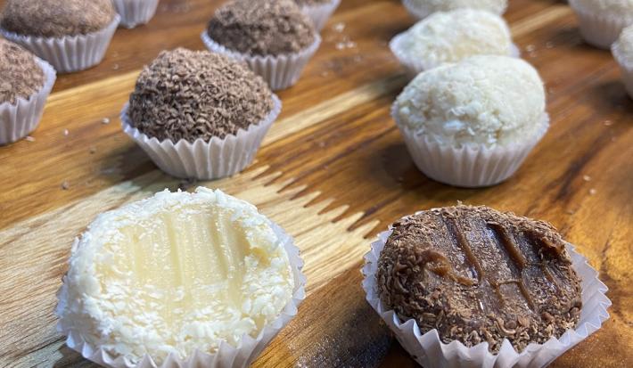 Fotografia em tons de marrom e branco em uma bancada de madeira de cor marrom. Ao centro, estão várias forminhas enfileiradas com brigadeiros de chocolate e brigadeiro branco.