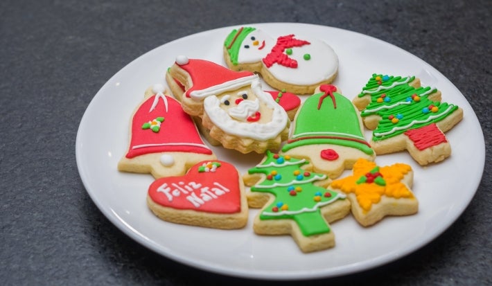 Em cima de uma mesa, prato branco com biscoitos de papai Noel, árvore de Natal e botas, decorados com glacê colorido e confeitos de bolinha coloridos.