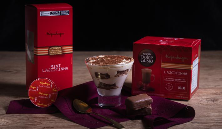 Fotografia em tons escuros de um recipiente de vidro com tiramisú sobre um pano vinho com uma colher de sopa e duas lajotinhas ao lado de uma embalagem de mini lajotinha e uma outra de dolce gusto lajotinha.