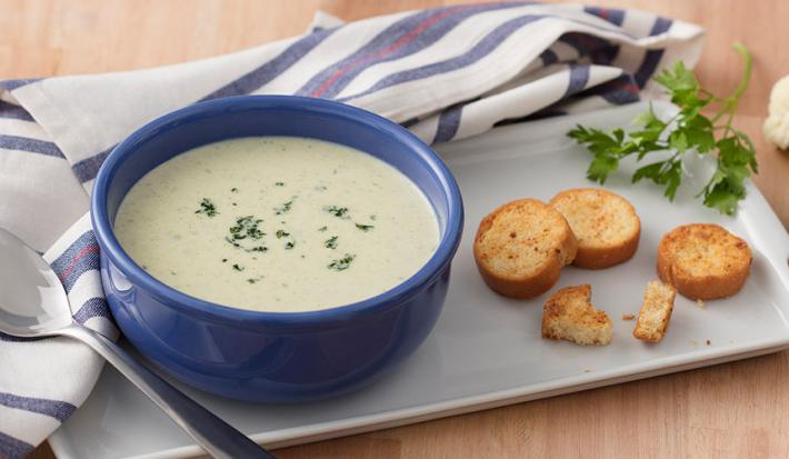 Fotografia em tons de azul e verde, com cumbuca azul ao centro com creme de legumes e salsa picada com torradinhas sobre travessa branca, ramo de salsa, brócolis e couve-flor decorando, guardanapo branco e azul com colher, sobre bancada de cor palha.