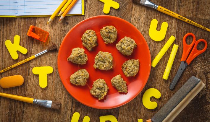 Fotografia em tons de laranja e amarelo em uma bancada de madeira escura, com objetos de itens escolares (tesoura, lápis de cor) e um prato laranja redondo ao centro com alguns cookies feitos com Mucilon.