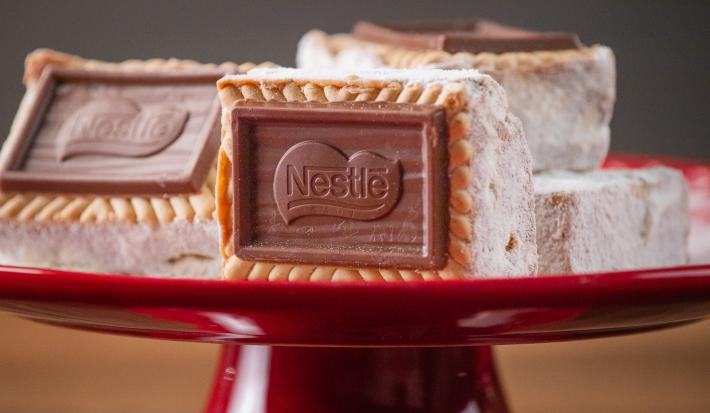 Fotografia de quatro unidades de palha italiana choco biscuit passadas no leite em pó. tem duas unidades no fundo, um por cima da outra, uma ao lado, e a palha italiana da frente está apoiada no centro da foto, todas estão sobre um apoio vermelho.