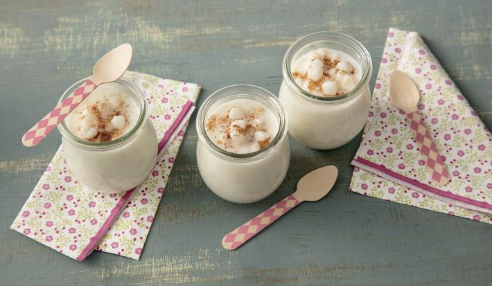 Foto de uma mesa de madeira cinza com 3 copinhos com mungunzá dentro e colheres e paninhos rosas decorando