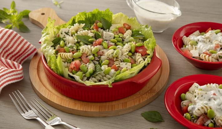 Fotografia em tons de verde em uma bancada de madeira de cor marrom. Ao centro, uma tábua de madeira contendo uma panela vermelha com a salada. Ao lado, há um garfo e um pano listrado vermelho. Ao fundo, há mini pratos com macarrão.
