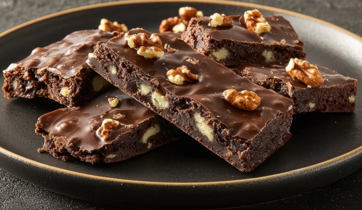 Em cima de uma mesa, barra com pedaços de massa com nozes, chocolate derretido e nozes picadas.