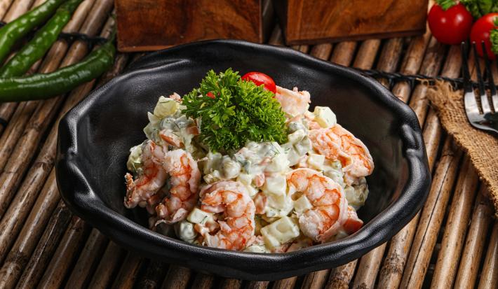 Fotografia de um recipiente preto fundo com uma salada de maionese com batata e camarão, com um tomate cereja e ramos de salsinha por cima. O recipiente está sobre uma mesa com madeiras desenhadas.