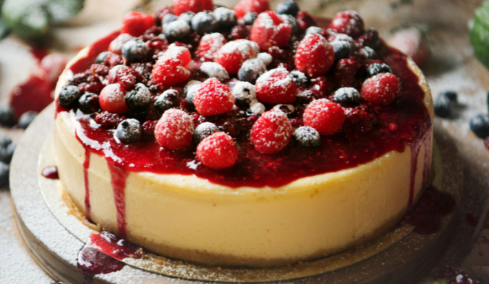Imagem de um cheesecake coberto com uma calda vermelha e frutas vermelhas. Está disposto sobre uma tábua de madeira redonda, em cima de uma mesa de mandeira.
