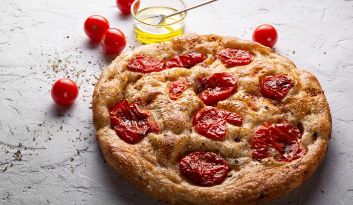 Fotografia de uma focaccia com tomate cereja assado, sal grosso e azeite. Ao lado da focaccia tem um recipiente de vidro pequeno com azeite, ao lado alguns tomates cereja, sobre uma mesa branca com texturas.