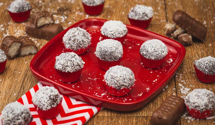 Imagem de uma prato vermelho com algumas unidades de Brigadeiro marrom passado em flocos de coco. A bancada está decorada com pedaços de bombons, um tecido vermelho e branco e mais brigadeiros.