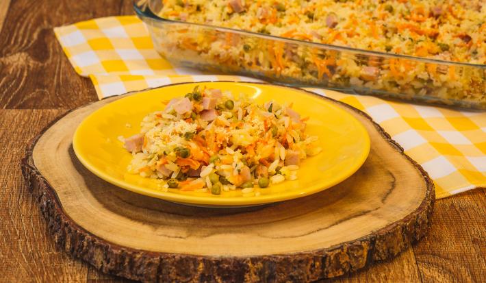 Foto de uma bancada de madeira. Sobre ela há uma toalha quadriculada amarela e branca e uma tábua de madeira com uma prato amarelo e a receita de arroz de forno. Ao fundo há uma travessa de vidro com mais arroz de forno