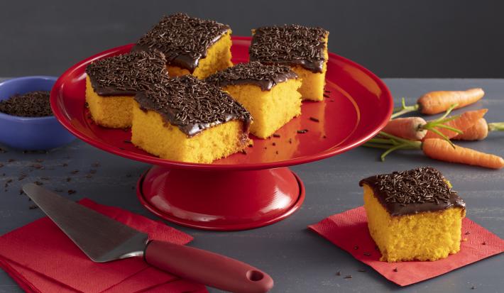 Fotografia em tons de vermelho e amarelo de uma bancada cinza, sobre ela um prato bailarina vermelho com 5 fatias de bolo de cenoura com cobertura de chocolate e granulado. Ao fundo mini cenouras.
