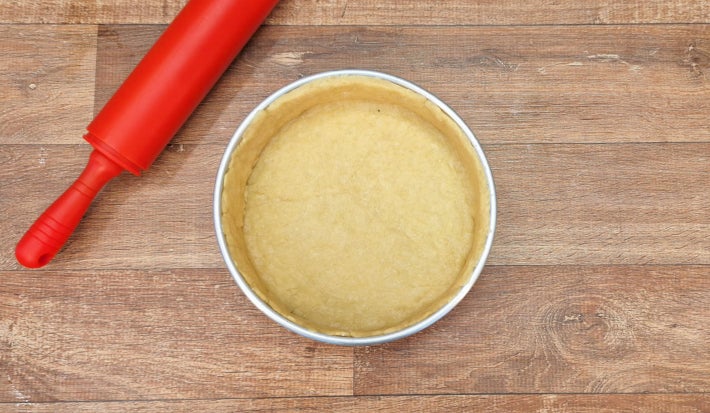 Massa forrando completamente o interior de uma forma redonda de alumínio ao lado de um rolo de cozinha vermelho.