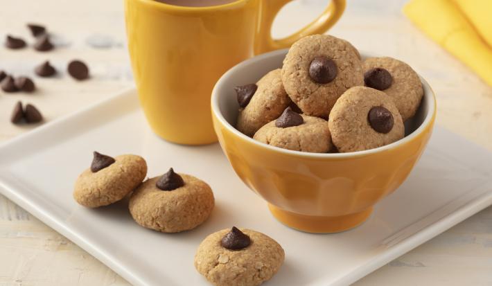 Fotografia em tons de branco e amarelo de uma bancada branca com um prato quadrado, sobre ele uma xícara amarela, um recipiente redondo amarelo com biscoitinhos  e três biscoitinhos. Ao fundo um paninho amarelo e gotas de chocolate.